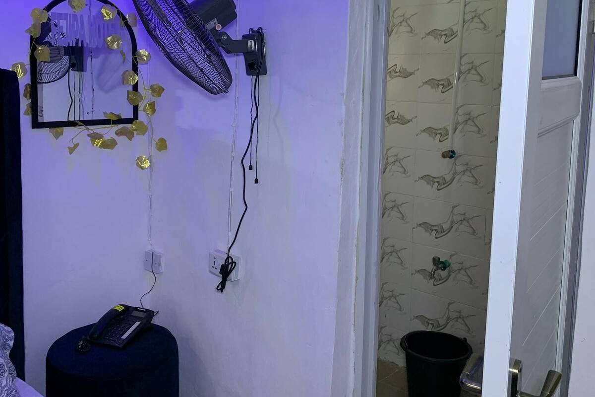 A room with a bed, a standing fan, a mirror, and a door leading to a bathroom with patterned tiles.