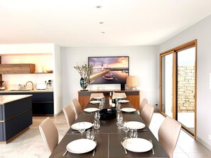 A dining area with a long table set for six, a kitchen with modern appliances, and a large window with a stone wall view. - Family Home, a 10-minute walk from the beach - 4 suites with private bathrooms (Quiberon)