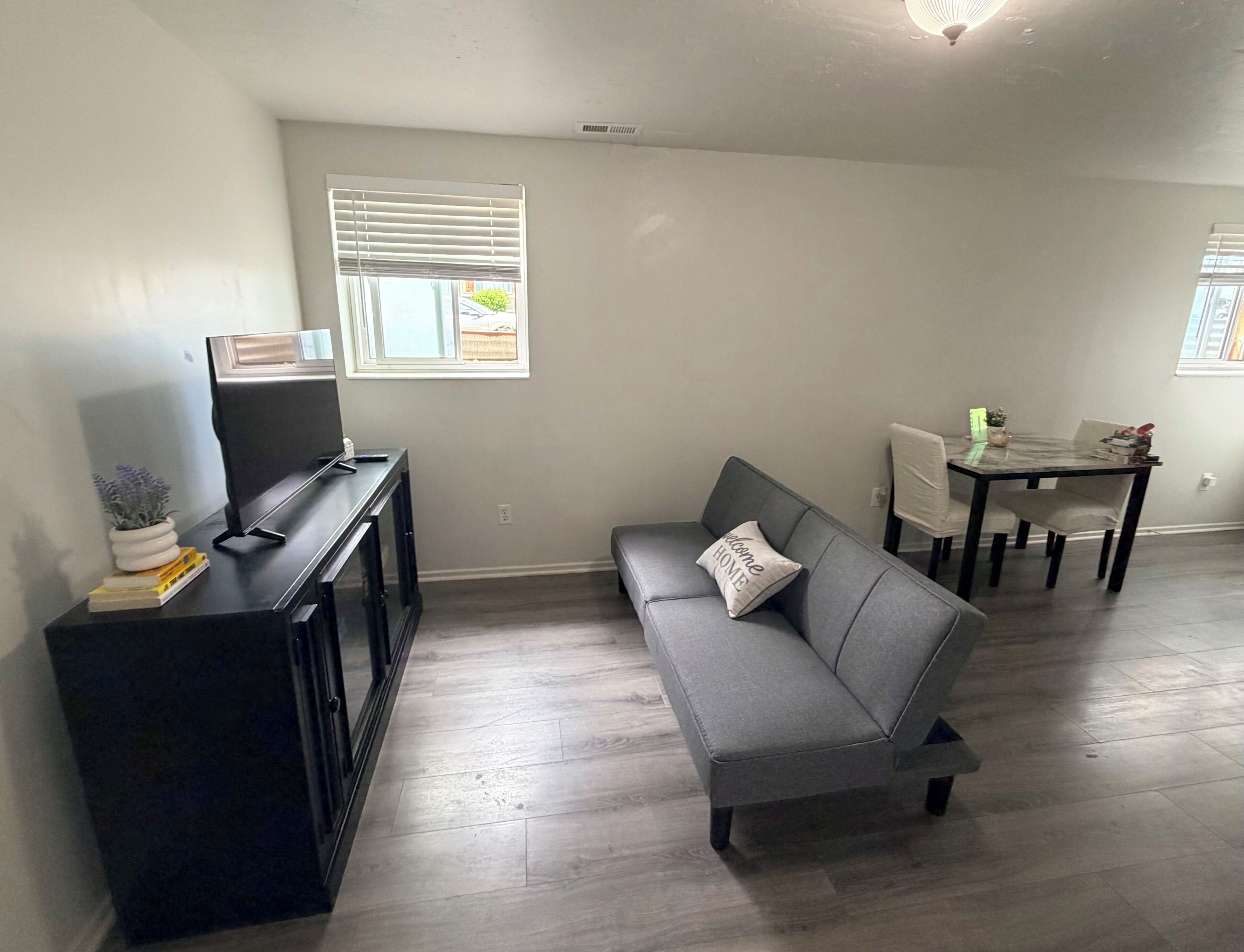 A living room with a grey sofa, a black entertainment unit, a dining table with chairs, and a window with blinds.
