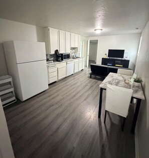 Cocina compacta con armarios blancos, un refrigerador, un microondas y una cafetera. Hay un comedor con una mesa y sillas, y un televisor al fondo.