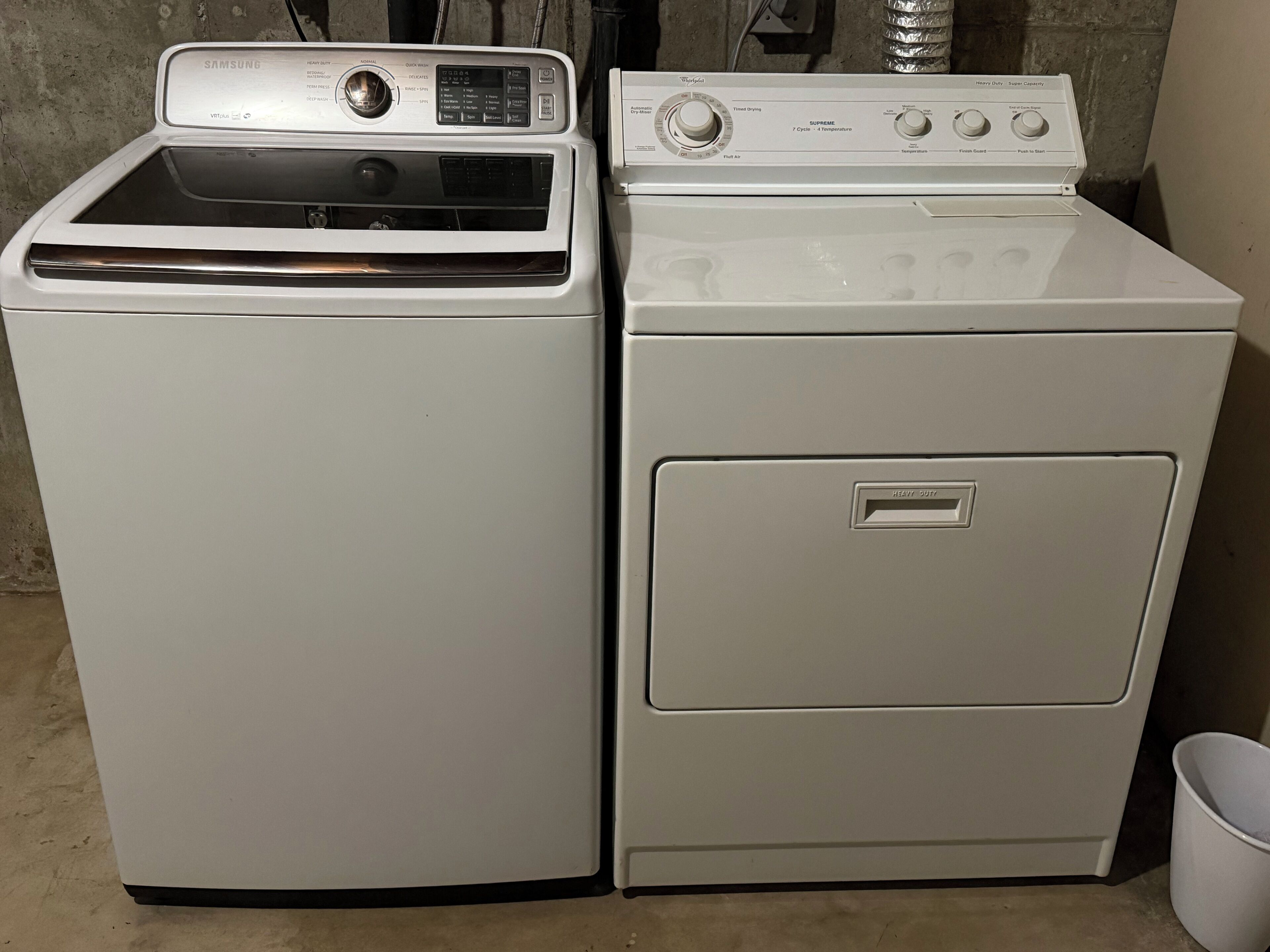 Twee witte wasmachines staan naast elkaar in een ruimte met een betonnen vloer en een metalen ventilatie rooster.