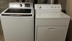 Deux machines à laver blanches, côte à côte, dans une pièce dont le sol est en béton et qui est équipée d’une grille de ventilation en métal.