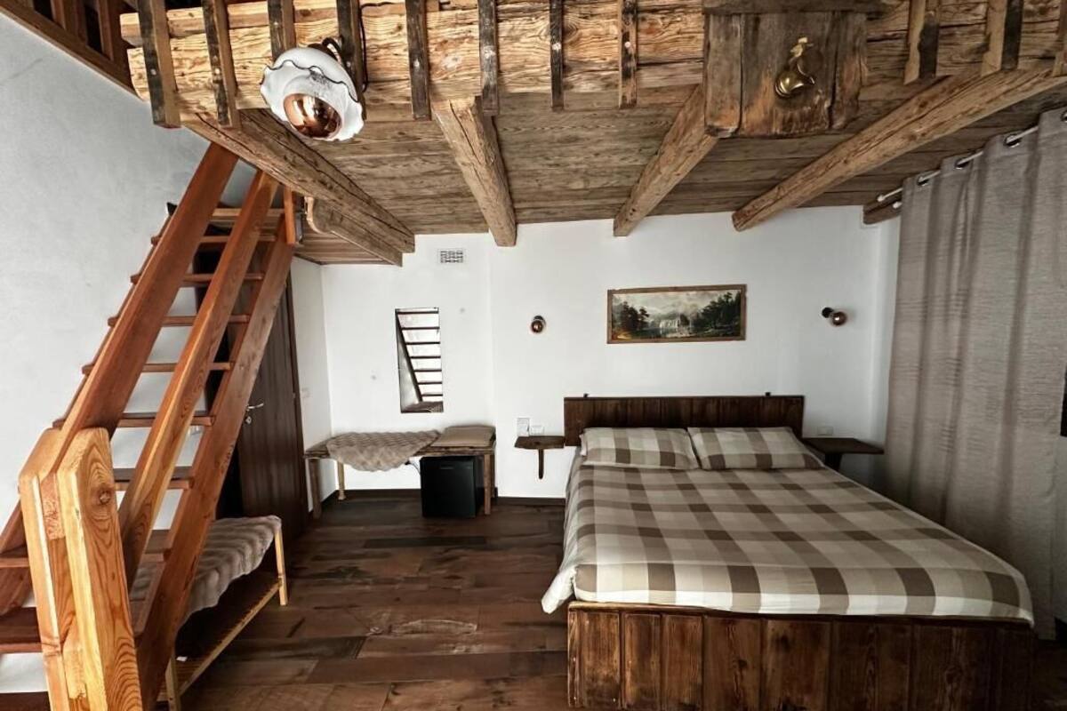 Un dormitorio con una cama de madera, una escalera de madera, un pequeño banco, un cuadro en la pared y un techo de madera.