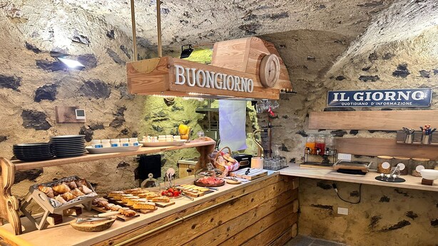 Una barra de desayuno rústica con estantes de madera, una máquina de café y un cartel que dice 'BUONGIORNO'.