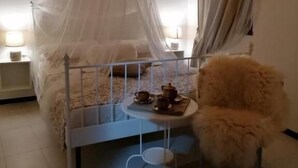 Un dormitorio con una cama con dosel, una pequeña mesa redonda con una silla, una mesita de noche con una lámpara y una ventana con cortinas.