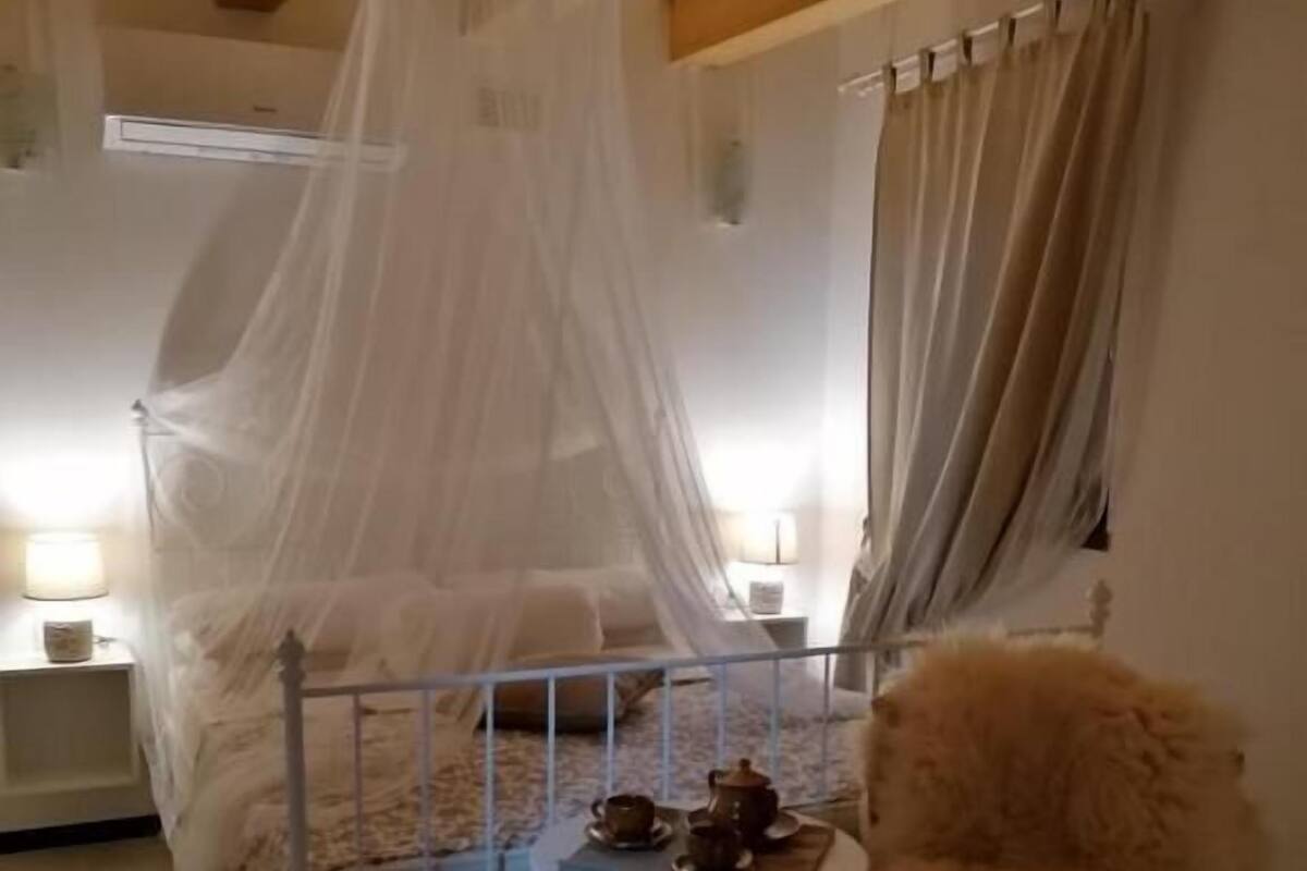 Un dormitorio con una cama con dosel, una pequeña mesa redonda con una silla, una mesita de noche con una lámpara y una ventana con cortinas.