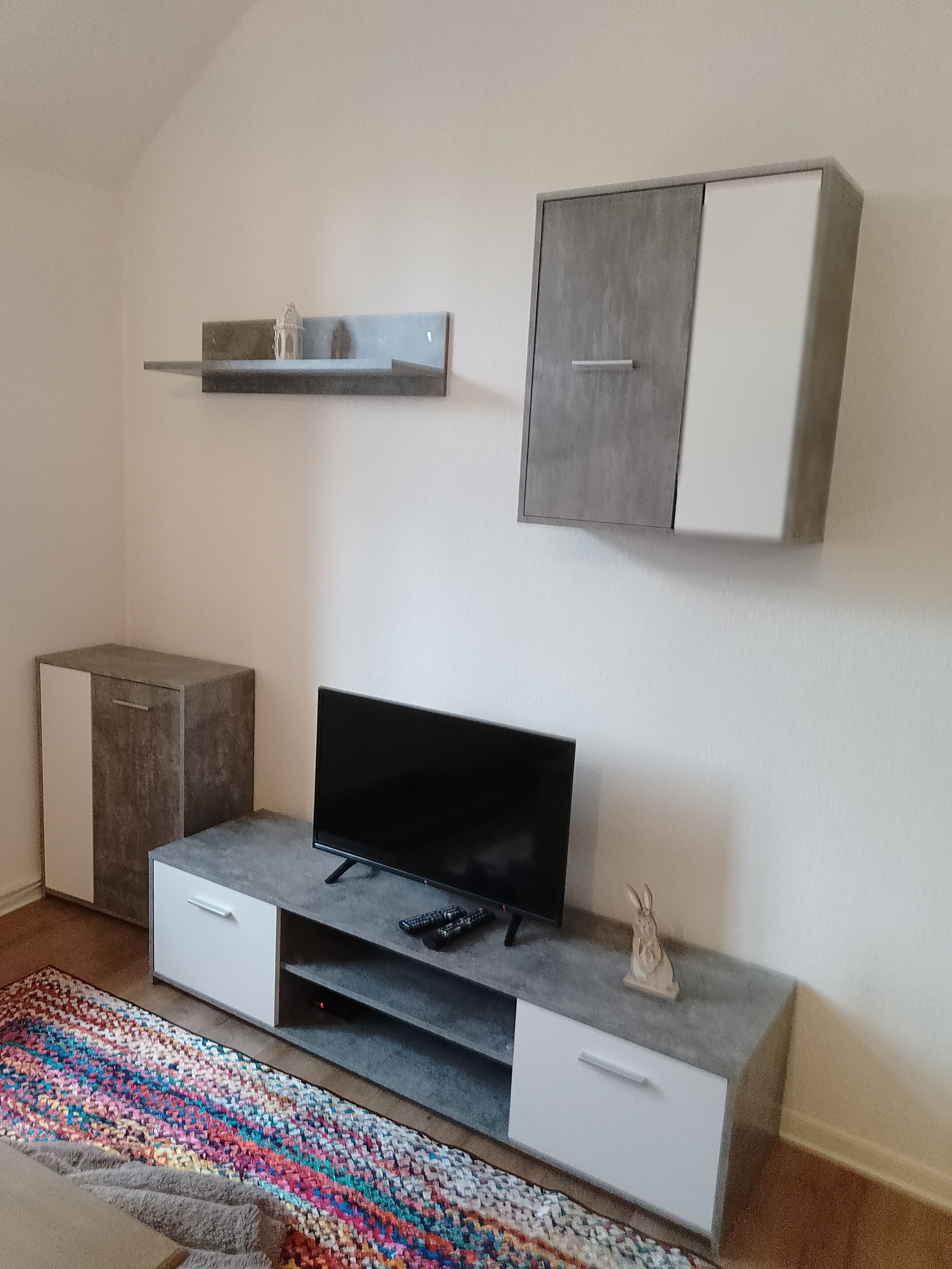 A modern living room with a flat-screen TV, grey cabinets, a small shelf, and a colorful rug.