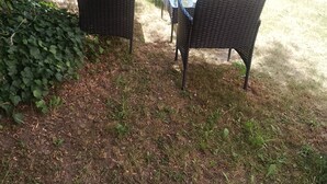 Un área de estar al aire libre con mobiliario de mimbre y una mesa de vidrio, rodeada de césped y una cerca de madera.