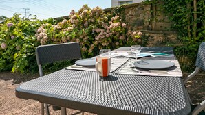 Et udendørs spisebord dækket med tallerkener, bestik og et glas vand, omgivet af frodigt grønt og en stenvæg.