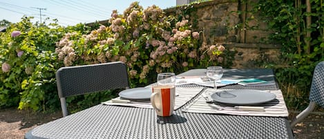 Mesa al aire libre dispuesta con platos, cubiertos y un vaso de agua, rodeada de abundante vegetación y un muro de piedra.