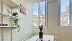 Ein Zimmer mit Esstisch, Stühlen, einem Heizkörper, Regalen mit Pflanzen und einem Fenster mit Blick auf Gebäude.