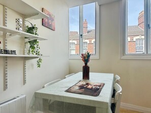 Ein Zimmer mit Esstisch, Stühlen, einem Heizkörper, Regalen mit Pflanzen und einem Fenster mit Blick auf Gebäude.