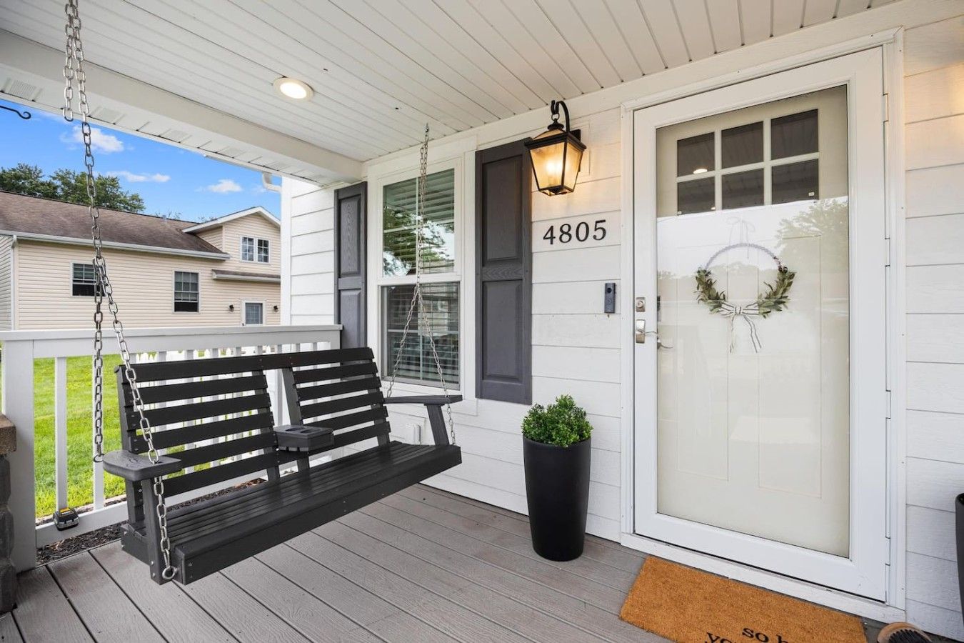 A porch with a black swing bench, a potted plant, a door with a number, and a wreath.