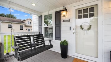 A porch with a black swing bench, a potted plant, a door with a number, and a wreath.