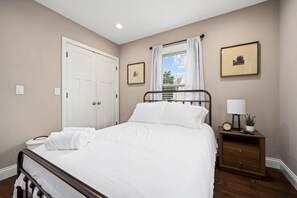 A bedroom with a bed, a nightstand, a window with curtains, a door, and framed pictures on the wall. - Peoria Heights Cottage: Arcade Fun on Columbus (Peoria Heights)