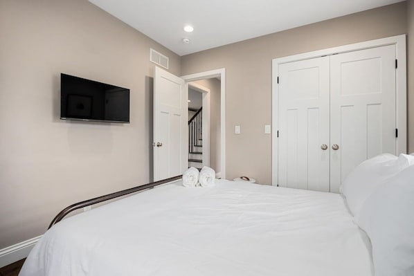 Quarto com cama, televisão fixada na parede e um conjunto de portas brancas.