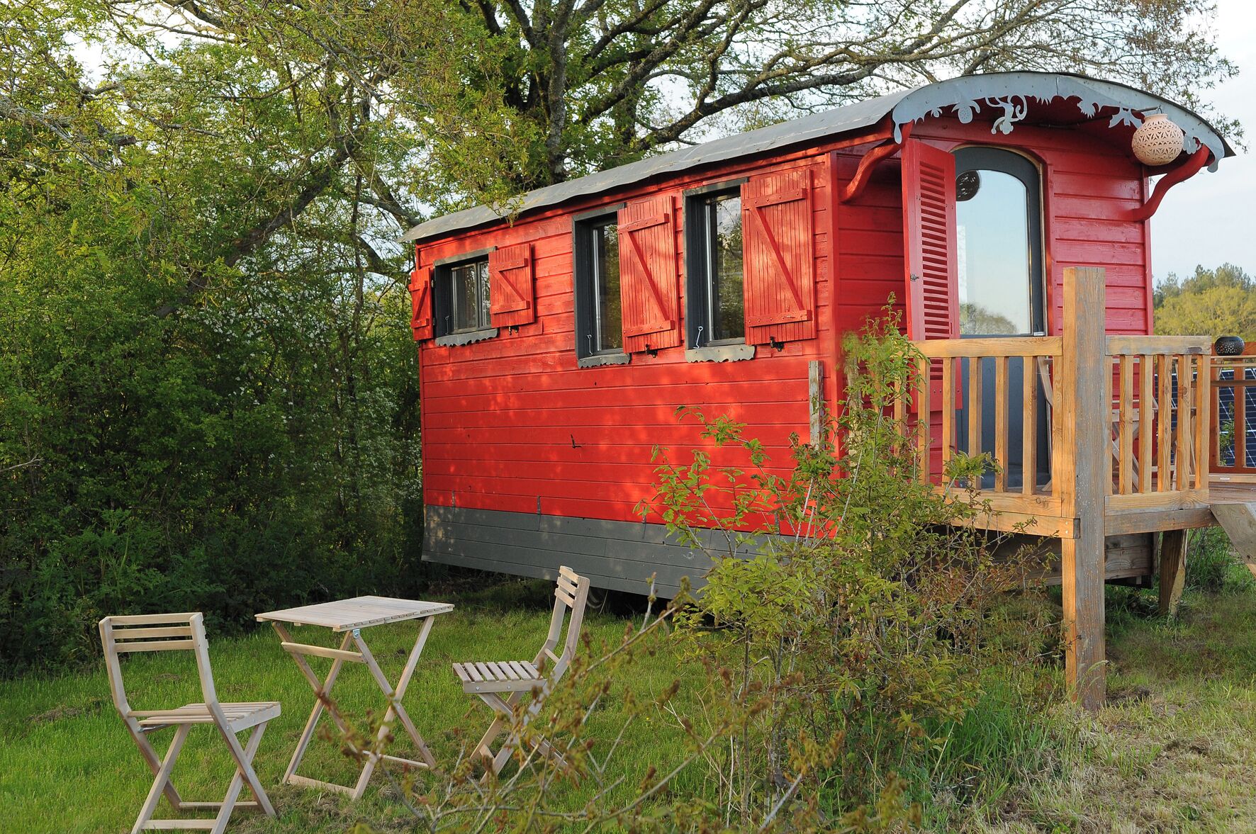 A red wooden cabin with a deck, surrounded by greenery.