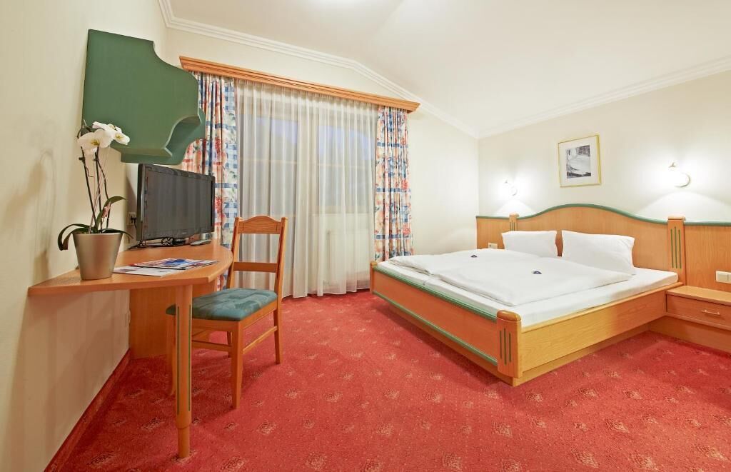A hotel room with a bed, a desk with a television, a chair, a window with curtains, and a nightstand.