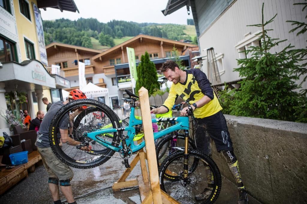A person washing a mountain bike in an outdoor setting with buildings and greenery in the background.