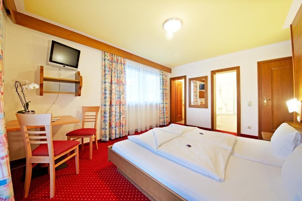Ett hotellrum med en säng, ett skrivbord med en stol, en tv som är monterad på väggen och ett fönster med gardiner.