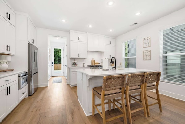 A modern kitchen with white cabinetry, a central island, and a dining area with wooden chairs. - Moxie - 1.5 Miles From DT & UofA (Fayetteville)