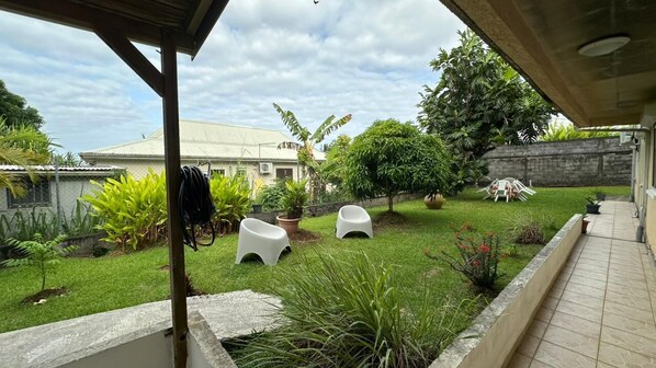 A covered outdoor area with a lawn, potted plants, and a white building in the background.