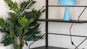 Une étagère métallique noire avec une lampe bleue sur l’étagère supérieure, une plante en pot et un magazine sur l’étagère du milieu.