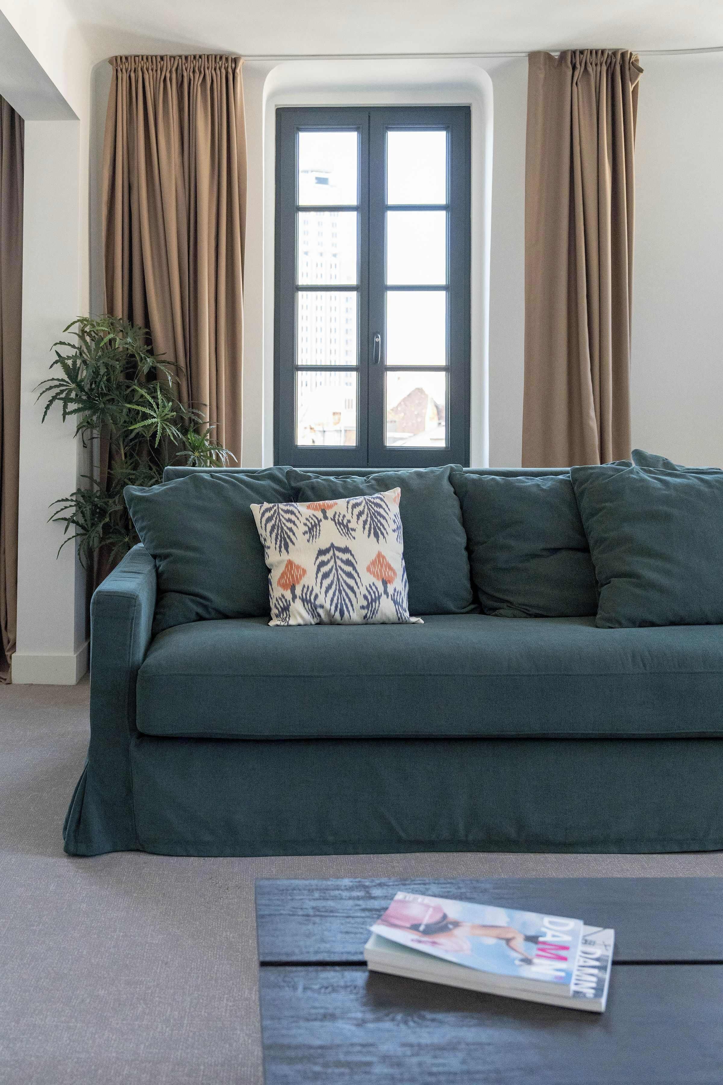 A living room with a dark green sofa, a patterned pillow, a magazine on a coffee table, and a large window with curtains.