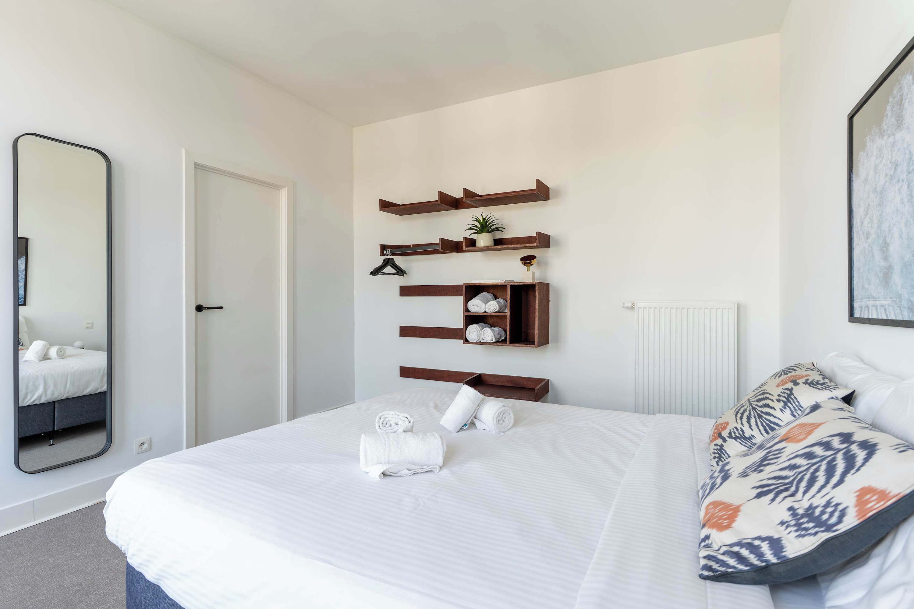 A neatly made bed with white linens, a large mirror, and a wall-mounted shelf with towels and a small plant.