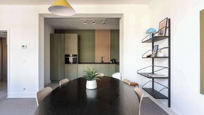 Ein moderner Essbereich mit einem runden Tisch, Stühlen, einer Zimmerpflanze, einer Pendelleuchte und einem schwarzen Metallregal mit Gegenständen.