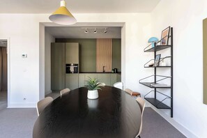 Uma sala de jantar moderna com uma mesa redonda, cadeiras, um vaso de planta, uma luminária pendente e uma prateleira de metal preto com objetos.