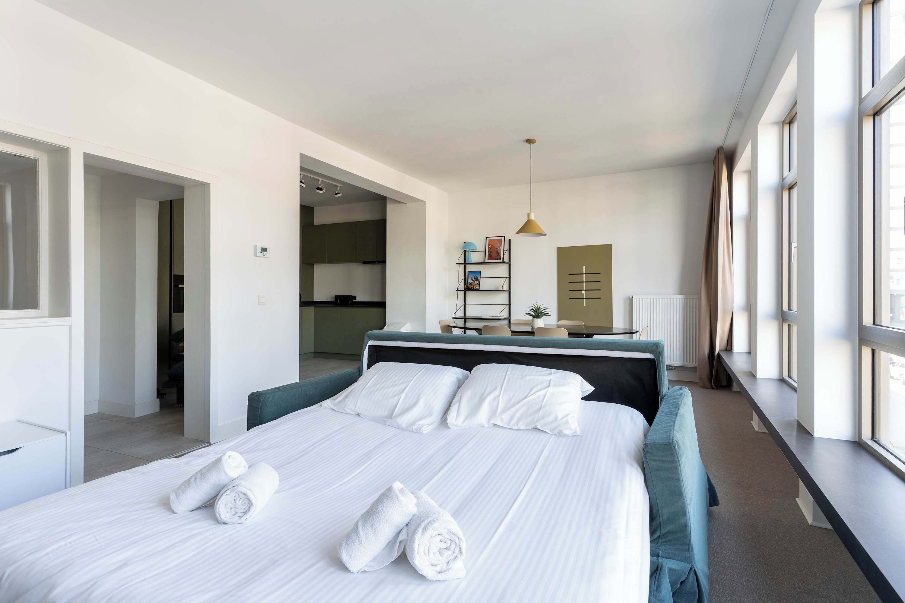 A modern hotel room with a large bed, a desk, and a view of the city through large windows.