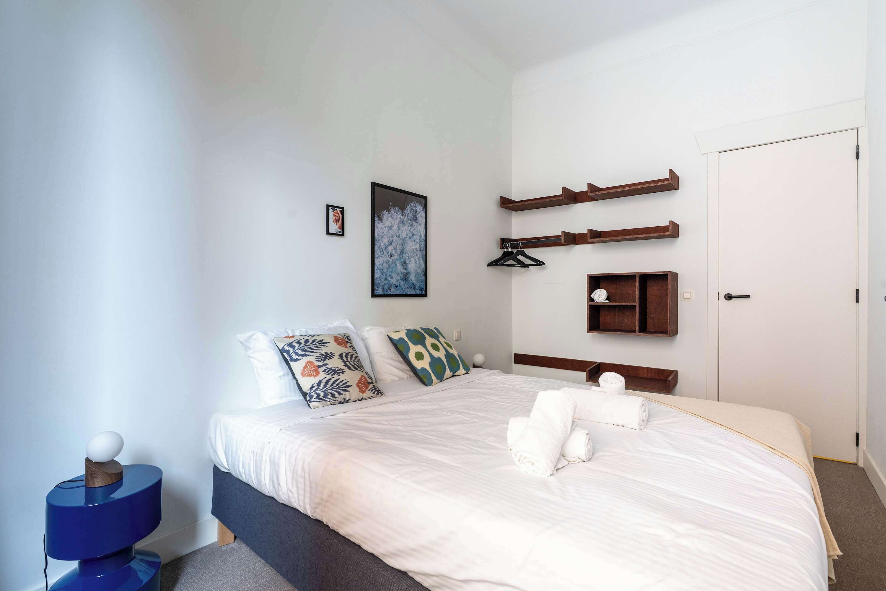 A bedroom with a bed, pillows, a bedside table, a wall-mounted shelf, and a door.
