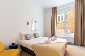 Quarto de hotel com cama, janela, radiador e uma pequena mesa com abajur.
