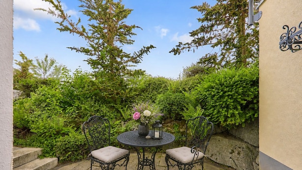 A patio with two chairs and a table, surrounded by lush greenery and a stone wall. - Villa Luna – Stylish Living. Arrive feeling relaxed. Feel at home. (Schaafheim)