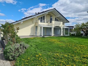 Ein modernes, zweistöckiges Haus mit großem Balkon, überdachter Terrasse und gepflegtem Rasen mit gelben Blumen.