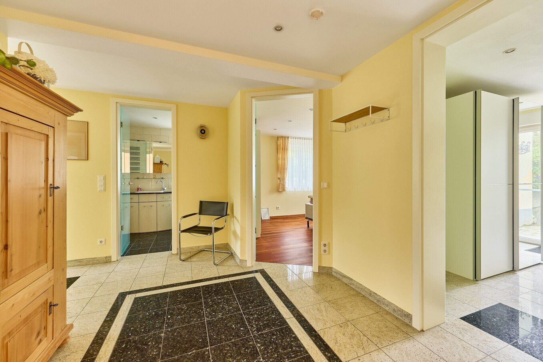 A hallway with a wooden cabinet, a chair, and a kitchen in the background.