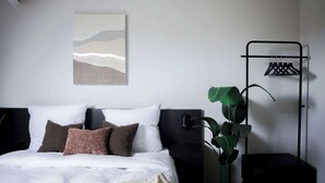 Ein Schlafzimmer mit Bett, Kissen, einem an der Wand hängenden Kunstwerk, einer Pflanze und einem Kleiderständer.