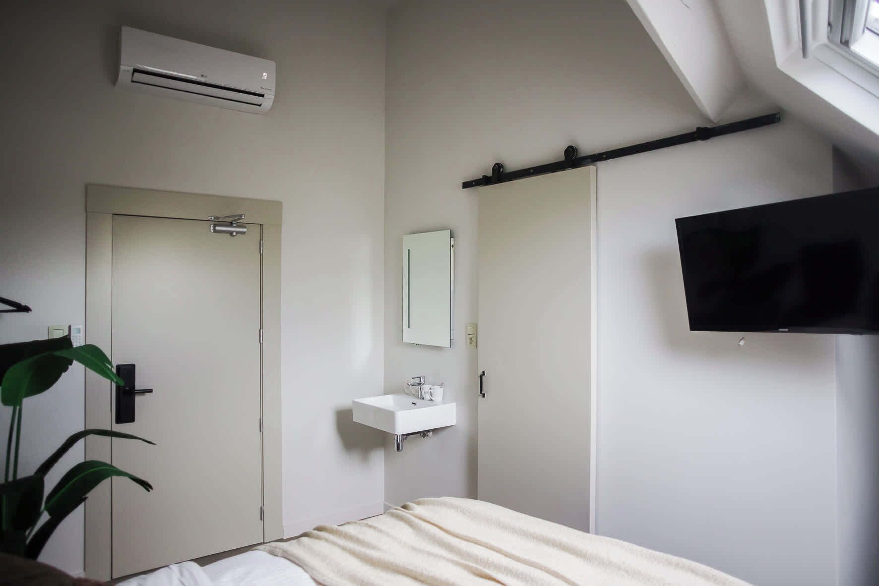A hotel room with a bed, a bedside table, a TV mounted on the wall, a white sink, and a door.