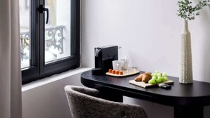 Ein Zimmer mit Fenster, Tisch mit Kaffeemaschine, Gläser, Teller mit Speisen, Vase mit Grünpflanzen und ein Stuhl.
