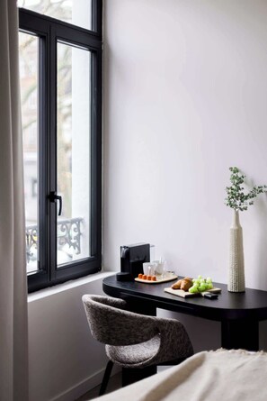 Et værelse med et vindue, et bord med kaffemaskine, glas, en tallerken med mad, en vase med grønne planter og en stol.