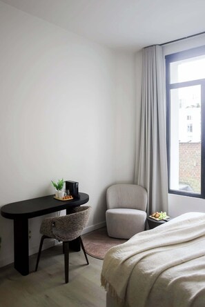 Habitació d'hotel amb un llit, una taula de treball amb una cadira, una taula petita amb una planta i una finestra amb cortines.