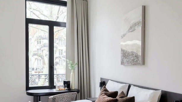 Ein Schlafzimmer mit einem Bett, einem Nachttisch, einem Fenster mit Vorhängen und einem an der Wand hängenden Kunstwerk.