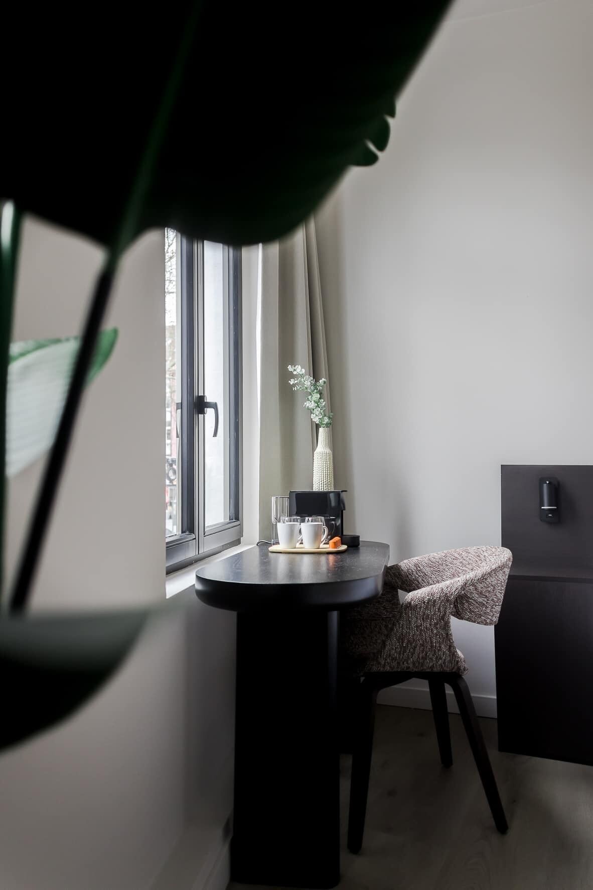 A small, modern room with a round table, a chair, a window with curtains, a potted plant, and a black cabinet.