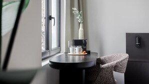 Ein kleiner, moderner Raum mit einem runden Tisch, einem Stuhl, einem Fenster mit Vorhängen, einer Topfpflanze und einem schwarzen Schrank.
