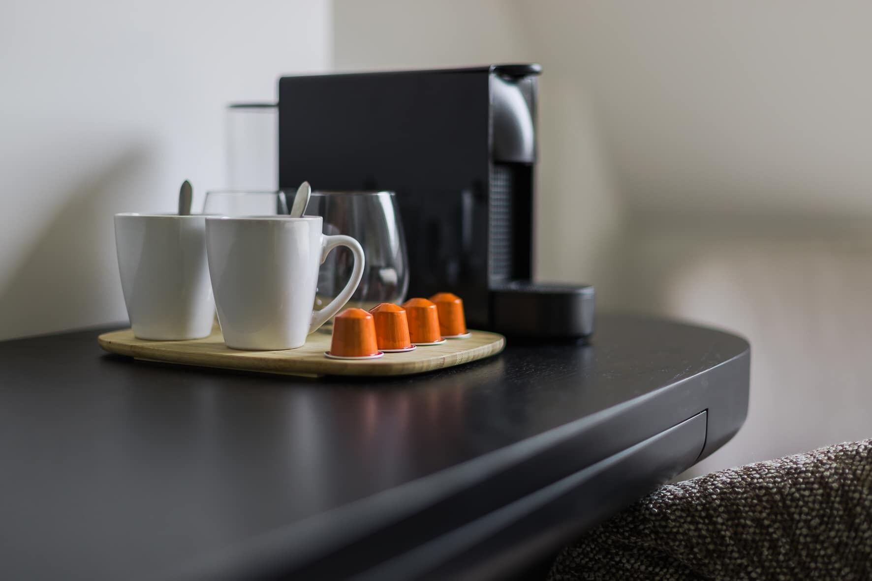 Una cafetera, dos tazas blancas y cápsulas anaranjadas sobre una mesa.