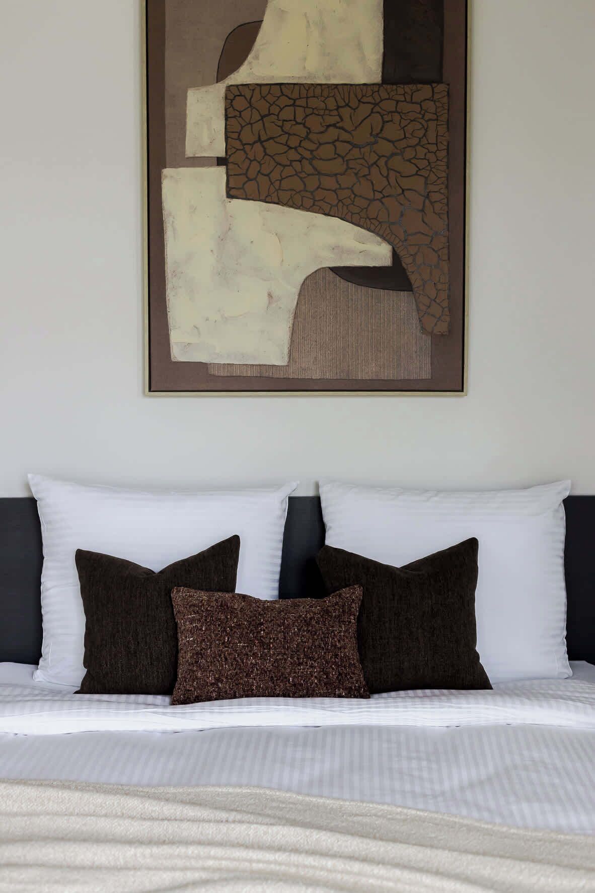 A bed with white and dark pillows, a textured pillow, and a framed abstract artwork above it.