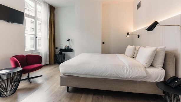 A modern hotel room with a large bed, a red chair, a small round table, and a desk with a lamp.