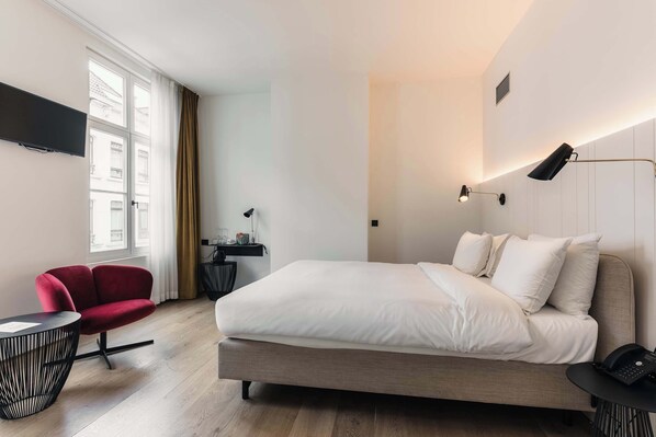 A modern hotel room with a large bed, a red chair, a small round table, and a desk with a lamp.