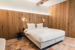 A bedroom with a wooden wall, a bed with white bedding, a small round table, and a black floor lamp.